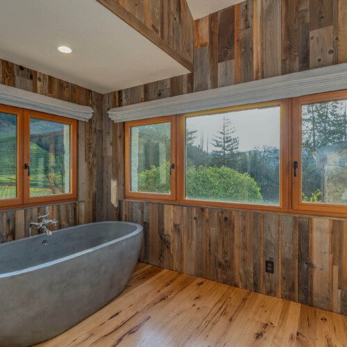 A large concrete soaking tub sits in a wood-paneled bathroom with views to vineyards