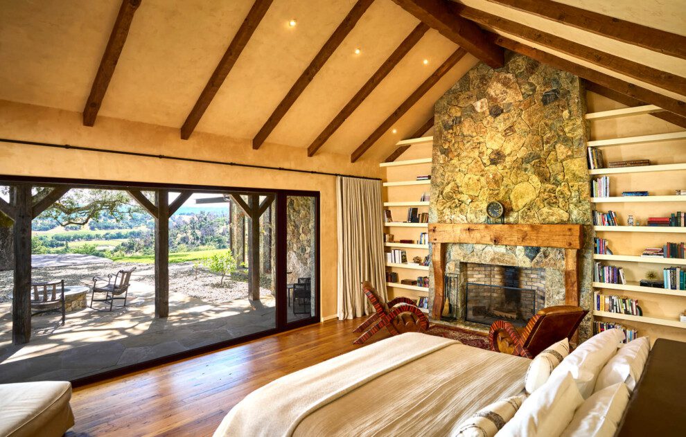 A large stone fireplace stands behind the king bed, which has a view out to a stone patio and hills beyond