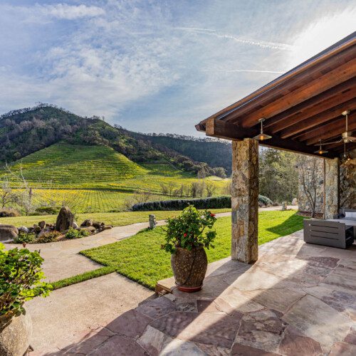 View past a stone patio with outdoor seating to rugged hills with vineyards